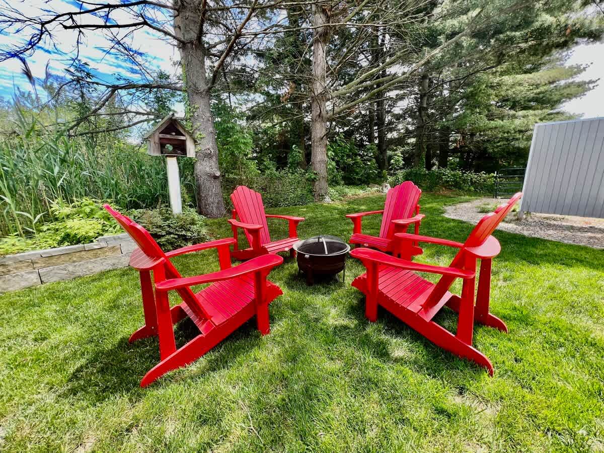 Outdoor fire pit area surrounded by four bright red Adirondack chairs on a lush green lawn, nestled among tall trees and natural foliage. A birdhouse on a post is visible in the background.