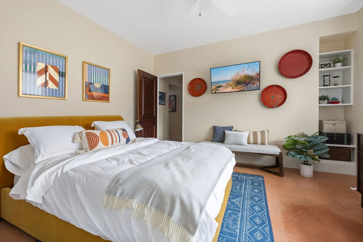 Spacious bedroom featuring a mustard yellow velvet bed, a long bench at the foot, and a wall-mounted TV. Decorative red plates and framed artwork adorn the light yellow walls.