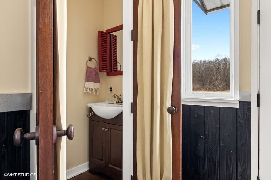 Cozy bathroom with a dark wood vanity, white sink, and a small window offering a view of trees. A red shuttered mirror cabinet hangs above the sink.