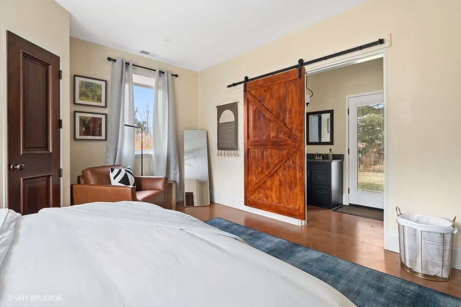 Bright bedroom with light yellow walls, a white bed, a brown leather armchair, and a large wooden barn door opening to a bathroom vanity area. A tall mirror leans against the wall.