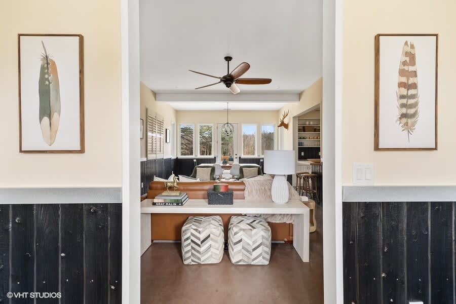 A welcoming view from a hallway with dark wood wainscoting and light walls, featuring two framed feather prints, looking into a bright living room with a ceiling fan, large windows, and a sofa.