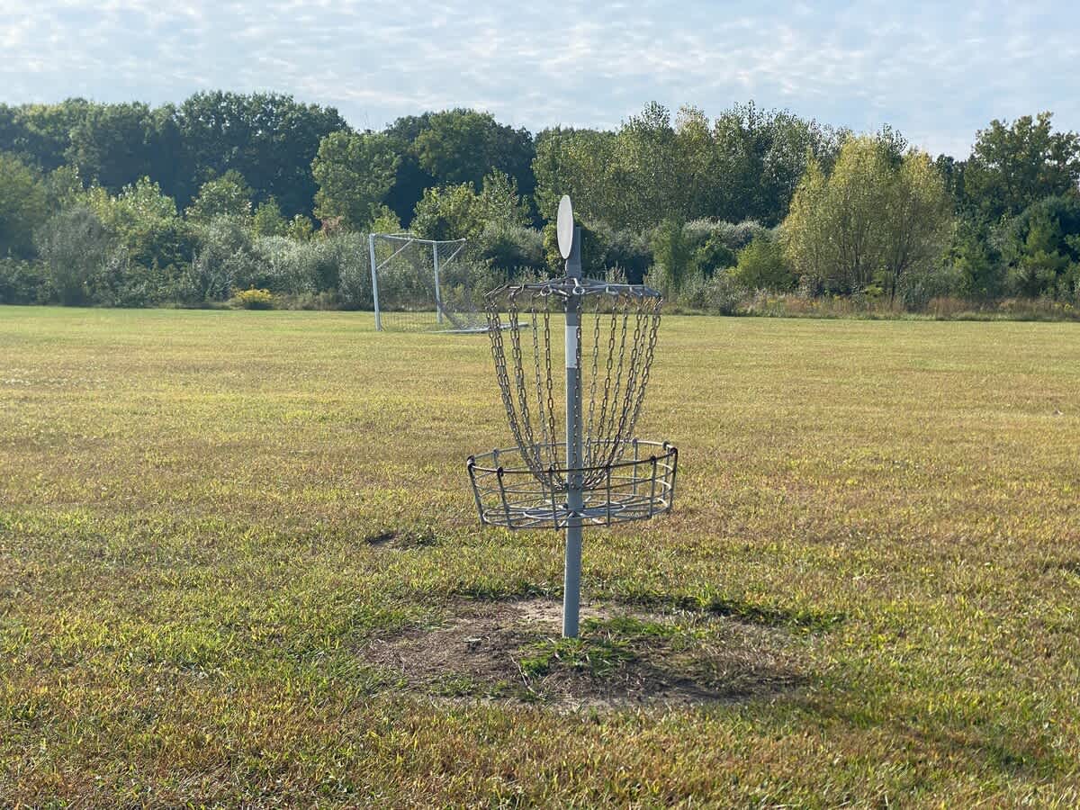 Metal disc golf basket with chains and a white target, positioned in a large, sun-drenched grassy field with a distant soccer goal and dense trees under a bright sky.