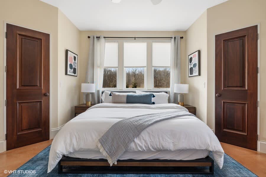 A bright bedroom with a queen-sized bed featuring white bedding and blue accents, flanked by two dark wood nightstands with lamps, and large windows overlooking the outdoors.