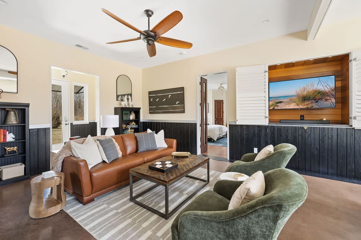 Spacious living room featuring a brown leather sofa, two green velvet armchairs, and a wooden coffee table on a striped rug. A large ceiling fan hangs overhead, and a wall-mounted TV is visible behind folding shutters.
