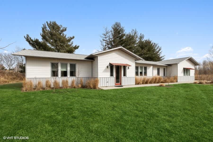 Modern single-story house with light-colored siding and multiple windows, set on a well-maintained green lawn. Tall trees and natural grasses surround the property.
