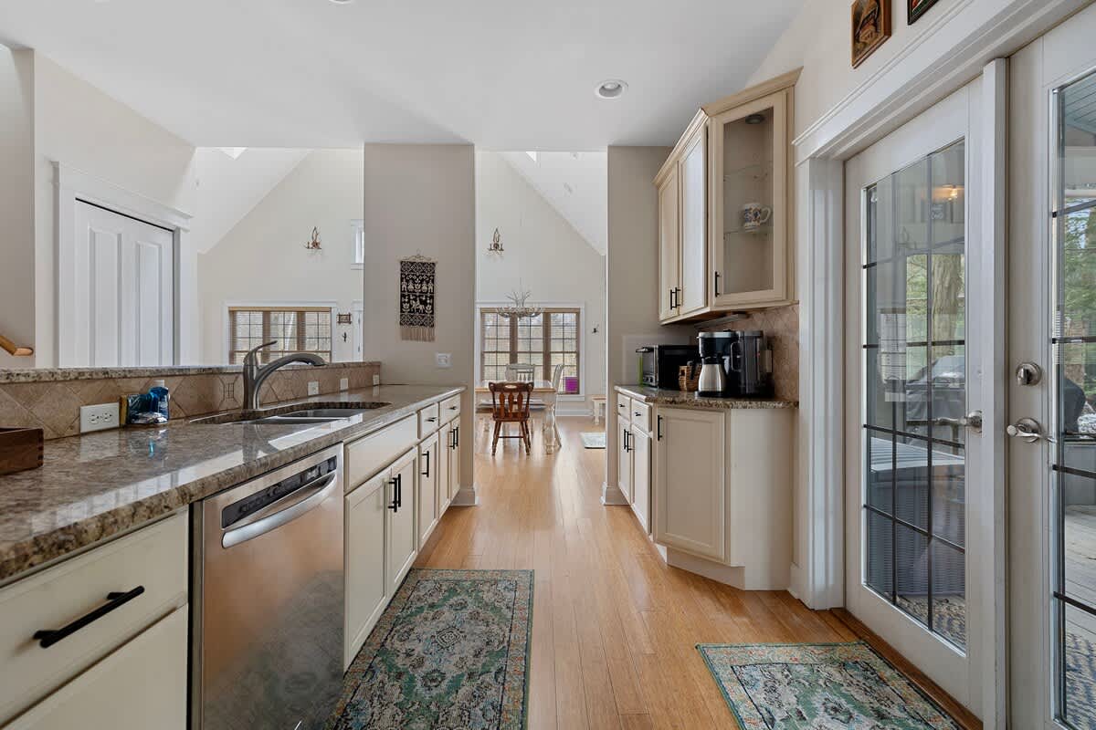 A bright kitchen with light wood flooring, granite countertops, and cream-colored cabinetry, featuring a stainless steel dishwasher and a view into a dining area with vaulted ceilings.