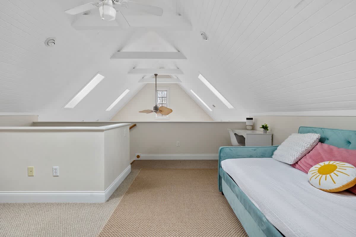 Bright attic loft with white paneled, sloped ceilings and skylights, featuring a teal daybed with colorful pillows, a small white desk, and light brown carpet. A ceiling fan hangs from the peak.