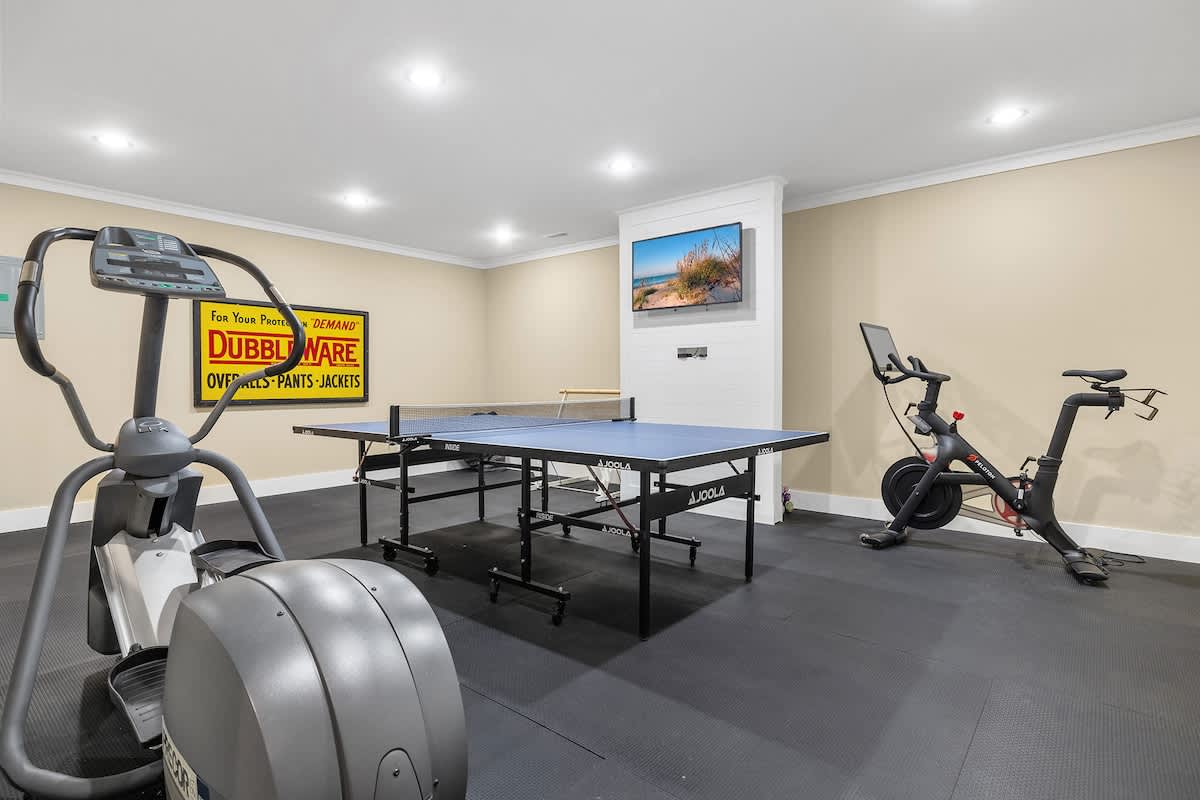A well-lit recreation room featuring a blue ping pong table, an elliptical machine, a Peloton bike, and a wall-mounted TV displaying a beach scene, all on a black rubber mat floor.