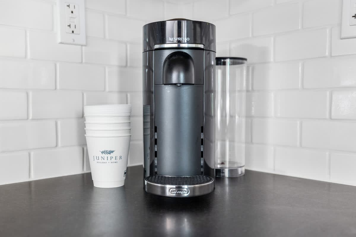 Close-up of a black Nespresso coffee machine and disposable cups on a dark kitchen countertop, set against a white subway tile backsplash.