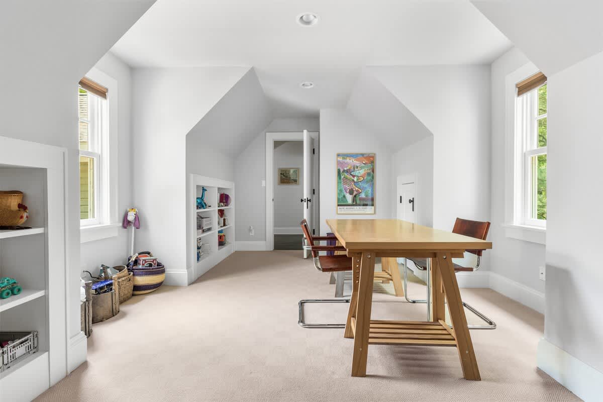 Bright attic room with sloped ceilings, light carpet, and white walls, featuring a large wooden table with two chairs, built-in shelves, and two windows providing natural light.