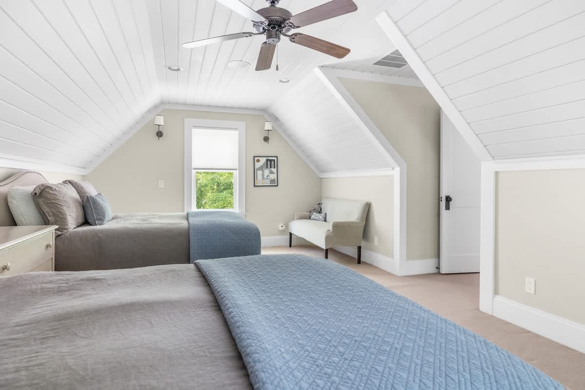 An attic bedroom with two twin beds, a sloped white shiplap ceiling, and a small upholstered bench, offering a comfortable and bright sleeping area.