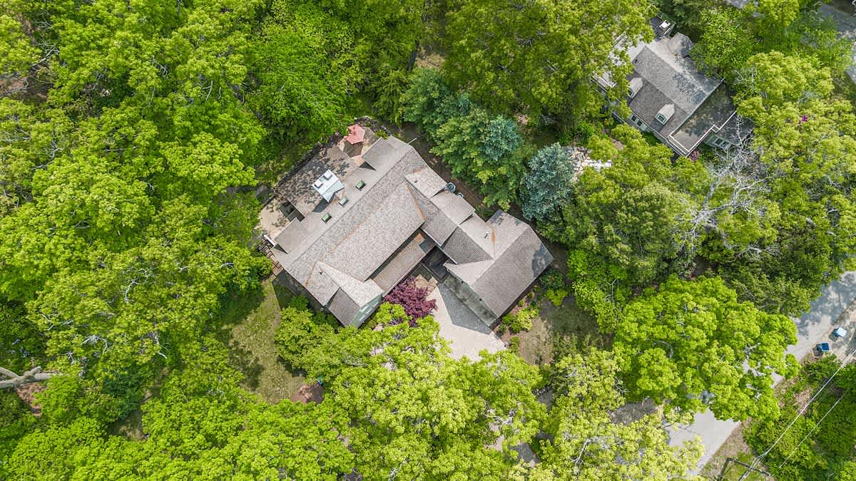 Aerial view of a sprawling house with a complex gray shingle roof, surrounded by a canopy of vibrant green trees. A paved driveway is visible, along with a deck area at the rear of the property.