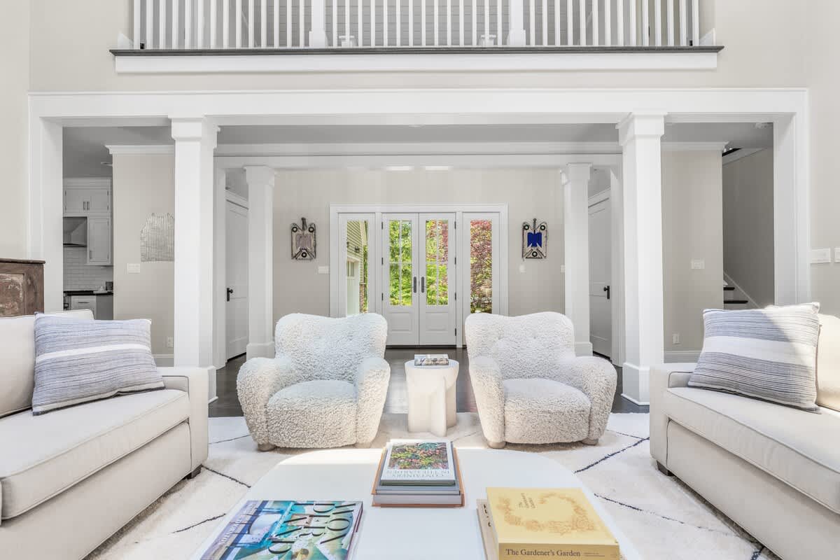 A grand living space with high ceilings and white columns, featuring two white sofas and two fluffy white armchairs arranged around a coffee table on a patterned rug. French doors lead to the exterior, and a balcony overlooks the space.