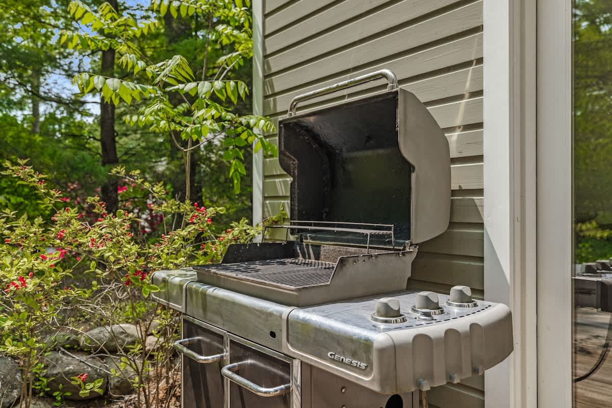 A close-up of a silver gas BBQ grill, positioned outdoors next to the house's olive green siding, surrounded by lush green foliage and flowering bushes.