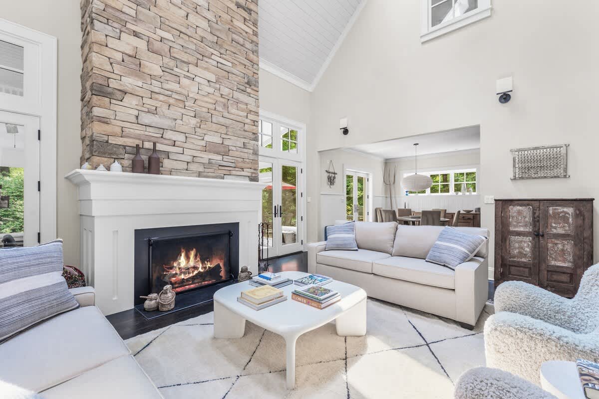 A bright and airy living room centered around a large stone fireplace with a white mantel. Two light-colored sofas and two fluffy armchairs are arranged on a geometric patterned rug, with a dining area visible in the background.