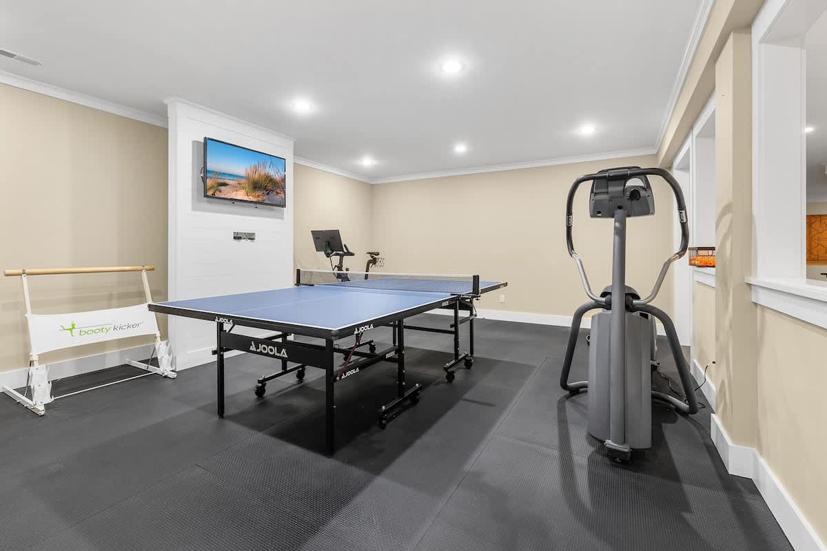 A spacious recreation room with dark rubber flooring features a blue ping pong table, an elliptical machine, and a stationary bike. A wall-mounted TV is visible above a white shiplap accent wall.