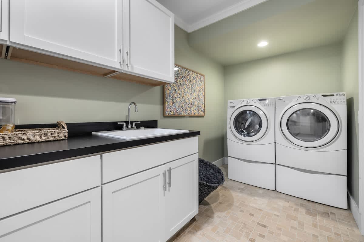 A functional laundry room with light green walls, white upper and lower cabinets, a black countertop with an integrated sink, and a side-by-side washer and dryer unit on pedestals.