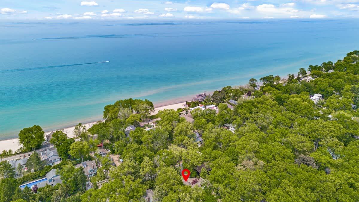 An aerial view showcases a lush green landscape with dense trees, a sandy beach, and the vast blue expanse of a lake or ocean. A red pin marks the location of the property amidst the greenery near the water.