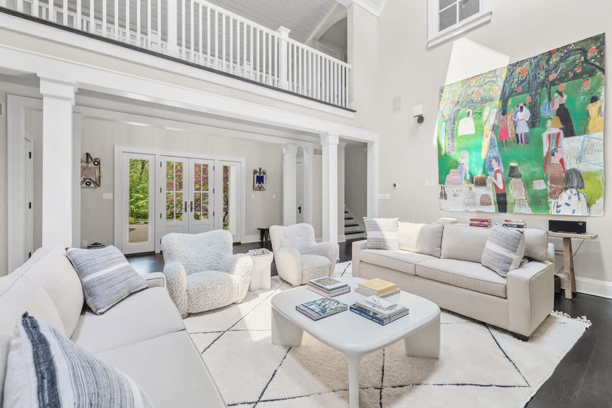 A spacious living room with high ceilings and white columns, featuring two light-colored sofas and two fluffy armchairs. A large, colorful abstract painting adorns one wall, adding an artistic touch to the bright space.