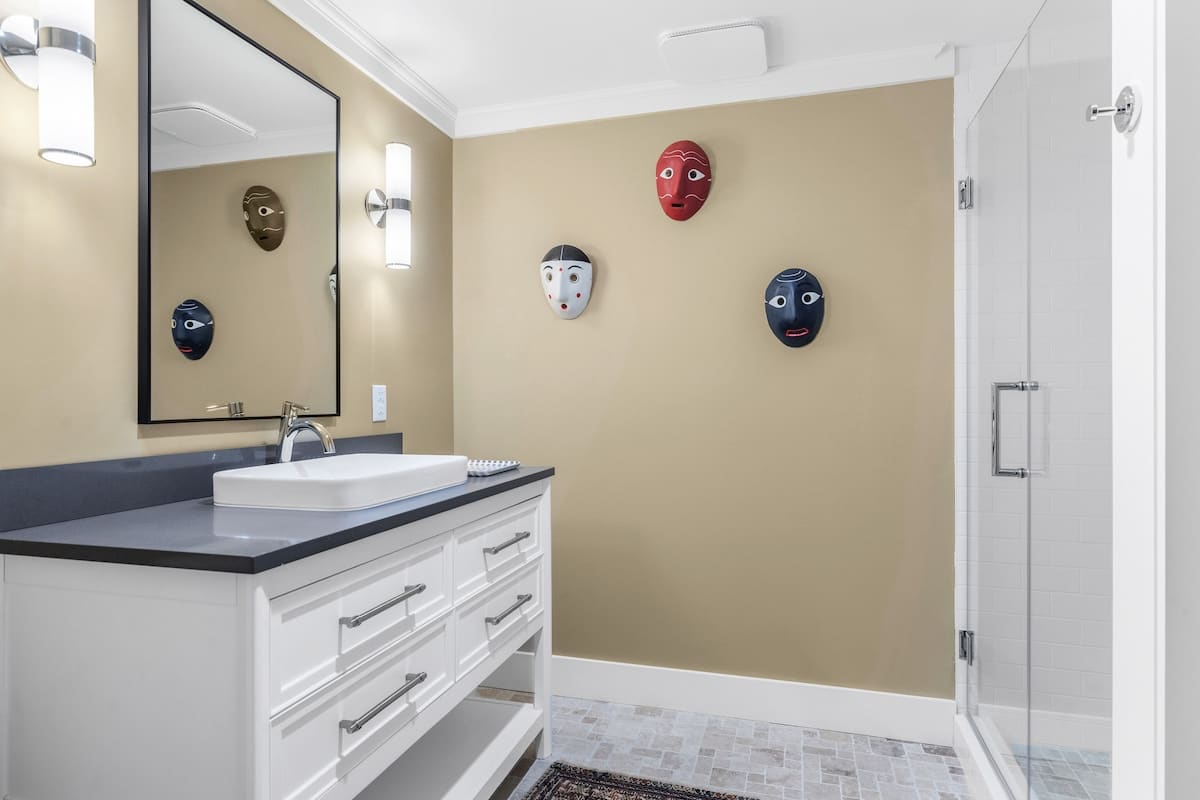 Modern bathroom with a white vanity, dark countertop, vessel sink, and walk-in glass shower, featuring light brown walls adorned with colorful decorative masks.