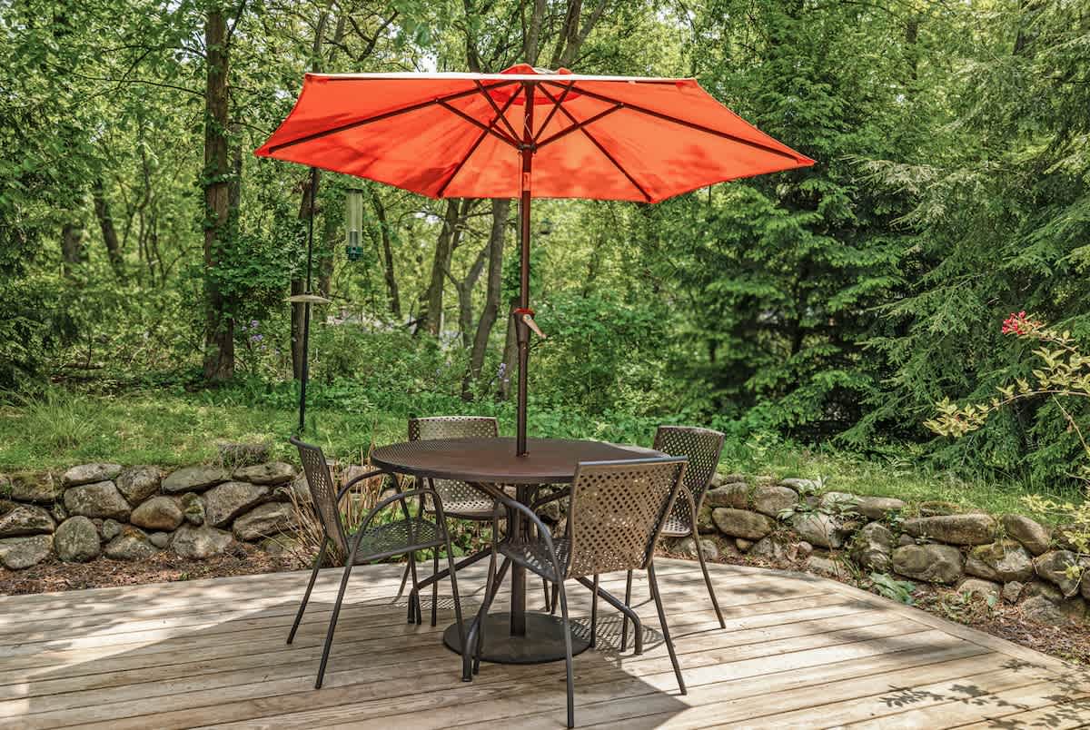 An outdoor dining area on a wooden deck, featuring a round table with four chairs and a bright orange umbrella, set against a backdrop of dense green trees and a stone retaining wall.