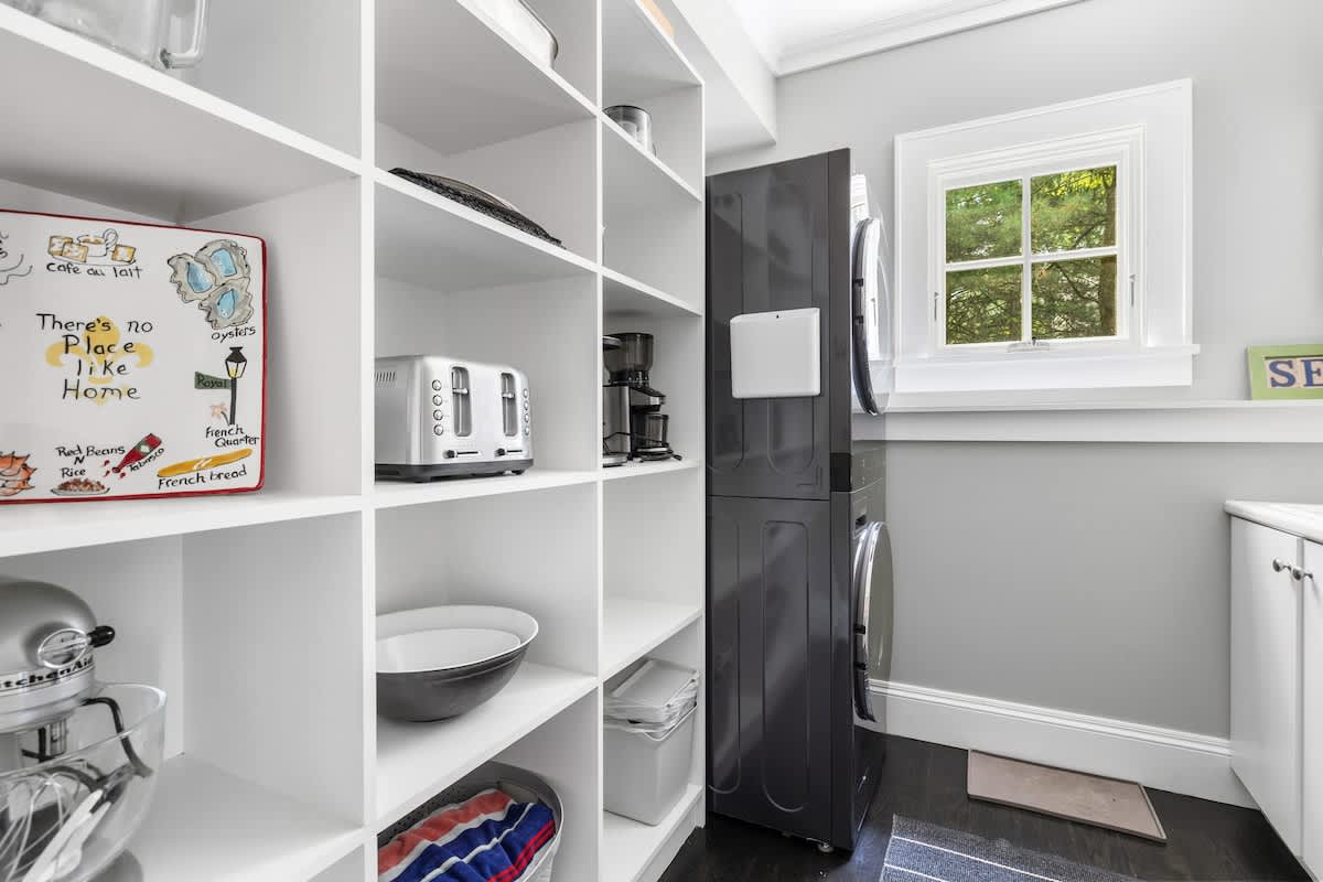 Organized pantry area with open white shelving stocked with kitchen appliances like a toaster and coffee maker, adjacent to a stacked black washer and dryer unit.