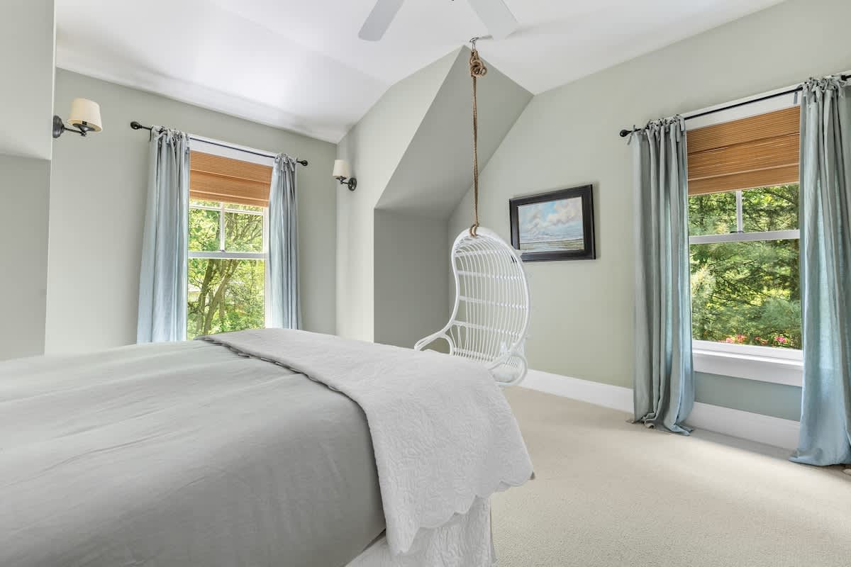 Serene bedroom with light green walls, light carpet, and a bed with light gray bedding, featuring two large windows with blue curtains and a unique white hanging chair.