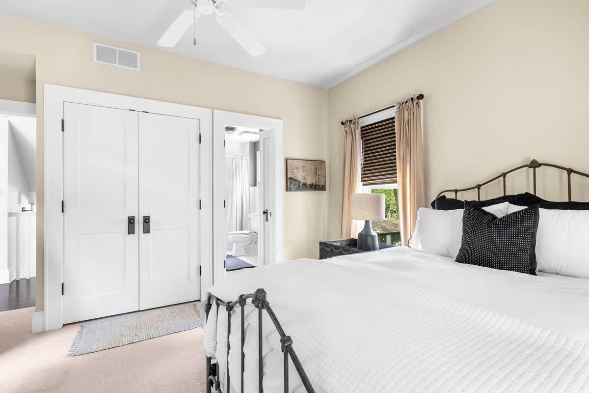 A bedroom featuring a black wrought iron bed, a double-door closet, and an open doorway leading to an en-suite bathroom, with a ceiling fan overhead.
