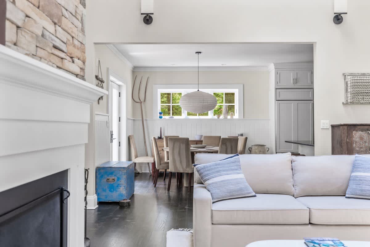 A view from the living room into an adjacent dining area, featuring a round wooden table with upholstered chairs and a unique pendant light. A glimpse of the kitchen with gray cabinetry is visible beyond.