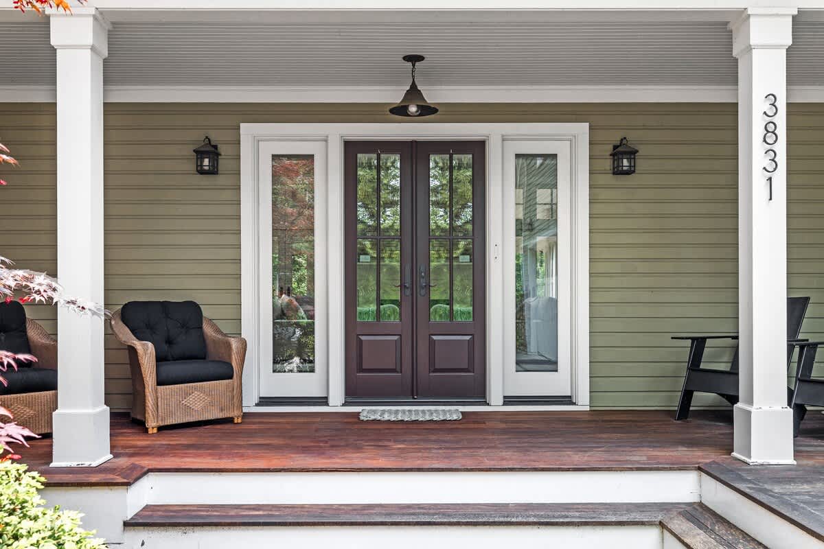 The inviting front porch of a house with olive green siding, featuring dark double entry doors flanked by glass sidelights, two wicker chairs, and the house number "3831" on a white column.