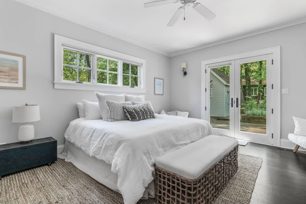 Serene bedroom featuring a large bed with white linens and a textured bench at the foot, light gray walls, a long window, and glass doors opening to an outdoor area.