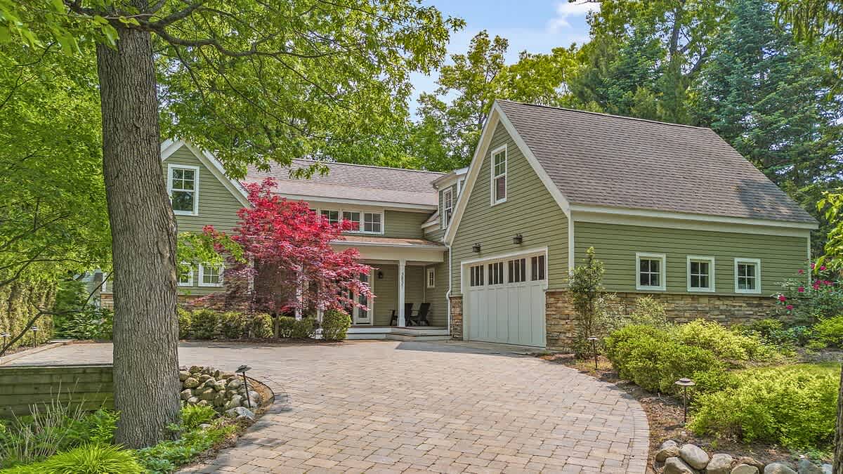 The front exterior of a large house with green siding and a stone foundation, featuring a paved driveway leading to a two-car garage, surrounded by mature trees and landscaping.
