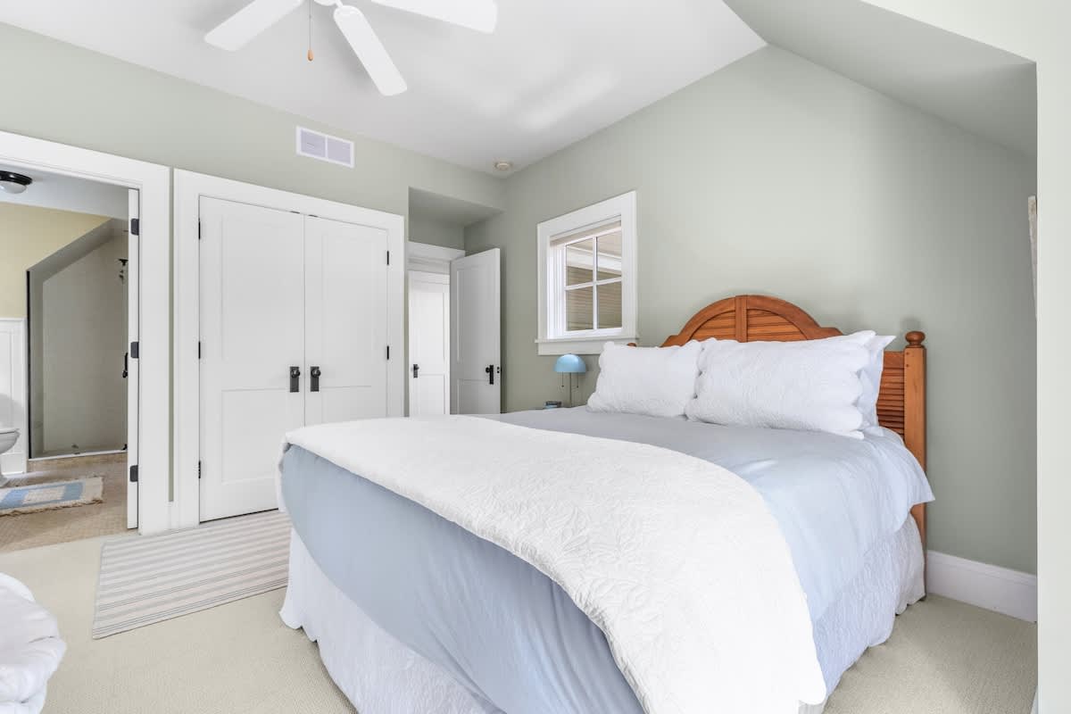 Cozy bedroom with light green walls, light carpet, and a wooden headboard bed with light blue bedding, featuring white double doors leading to a bathroom and a window.