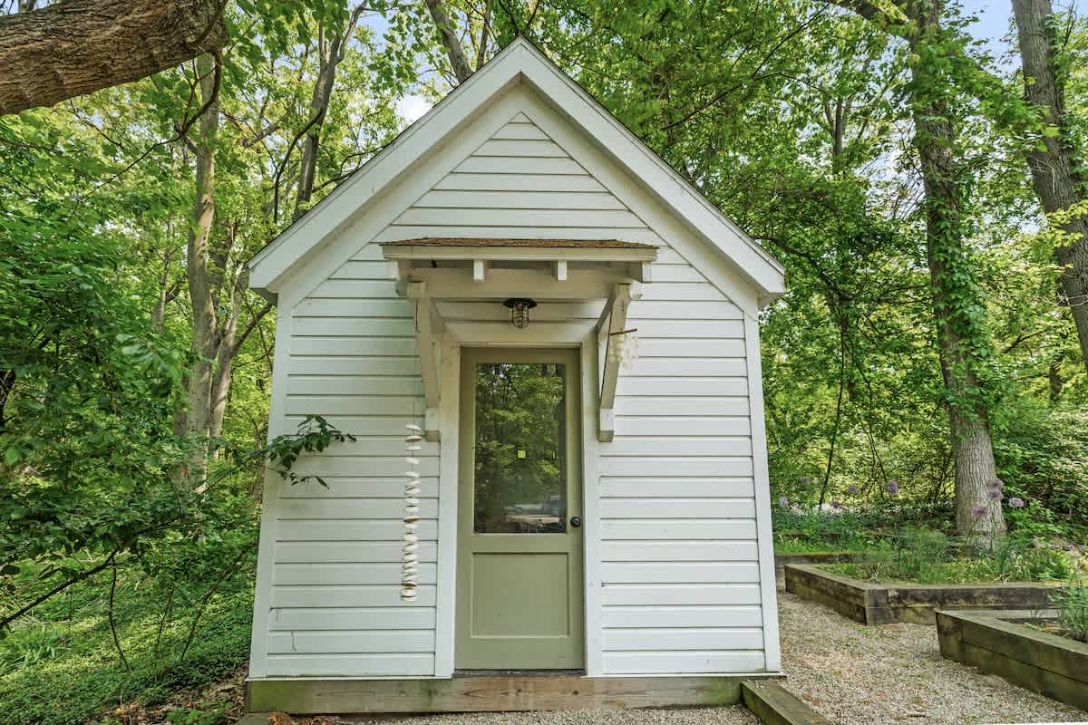 A charming small white shed with a green door and a small overhang, nestled among lush green trees and garden beds, suggesting a potting shed or garden retreat.