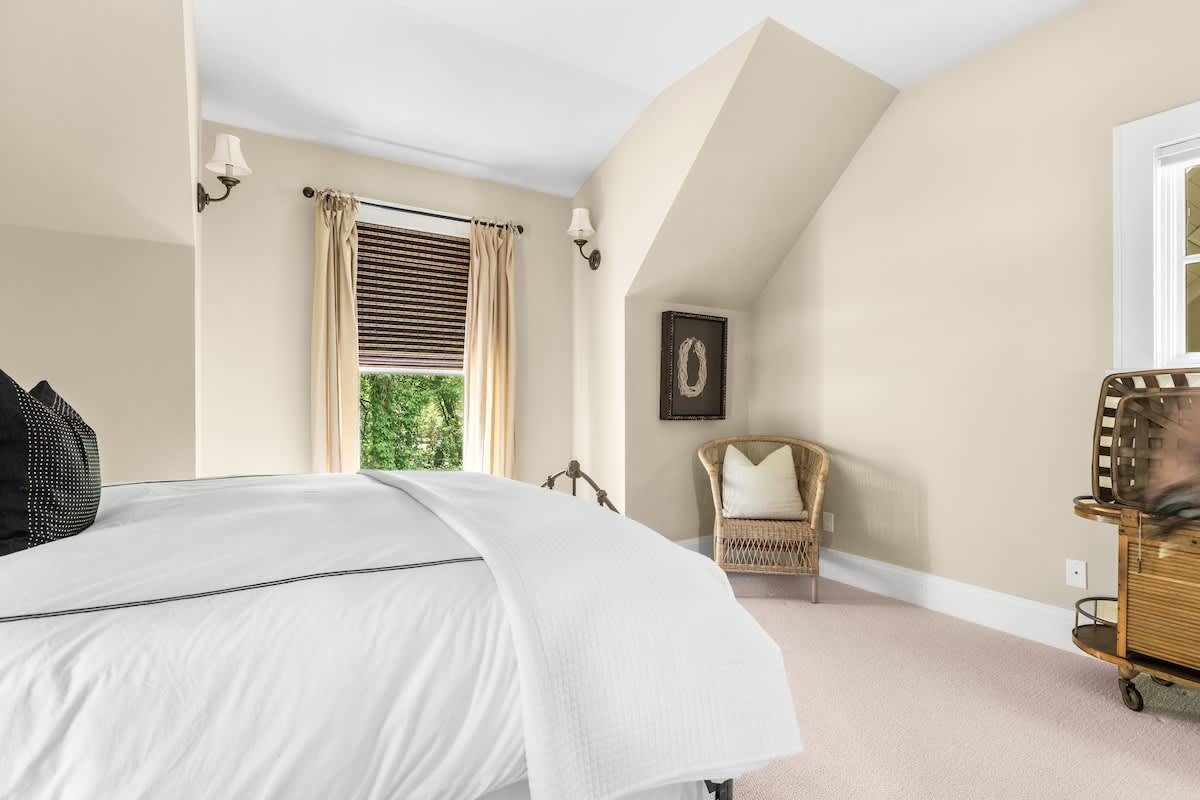 A corner view of the bedroom showing a black wrought iron bed, a wicker chair with a pillow, and a small side table, with light beige walls and carpeted flooring.