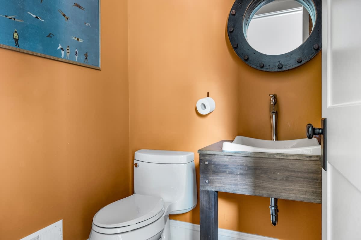 A compact powder room with vibrant orange walls, featuring a white toilet, a rustic wooden vanity with a white vessel sink, and a unique round porthole-style mirror above.