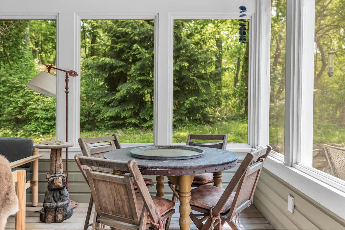 Cozy screened porch dining area featuring a rustic round wooden table with four chairs, surrounded by large windows offering expansive views of a dense green forest.