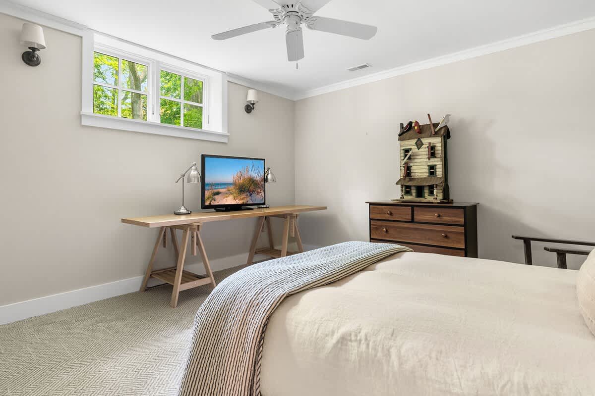 Neutral-toned bedroom with light beige walls and patterned carpet, featuring a bed, a wooden dresser with decor, a TV on a wooden console, and a window providing natural light.