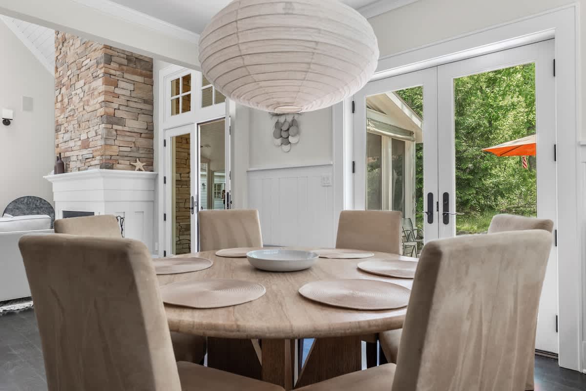 Bright dining area featuring a round wooden table with six upholstered chairs, illuminated by a large paper lantern, adjacent to a stone fireplace and glass doors opening to the outdoors.