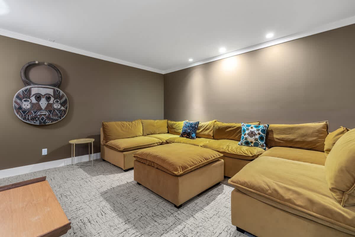 Spacious media room with dark brown walls and patterned carpet, featuring a large mustard yellow sectional sofa with decorative pillows and unique circular wall art.