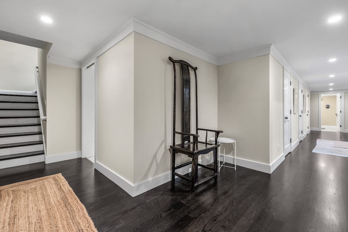 A spacious hallway or foyer with dark hardwood floors, light beige walls, and white crown molding, featuring a tall, dark wooden chair and a staircase with dark treads and white risers.