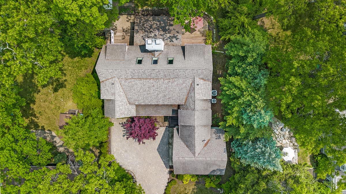 Overhead view of a large, multi-gabled house with a gray shingle roof, surrounded by lush green trees. A paved driveway leads to the front, and a deck with outdoor furniture is visible at the back.