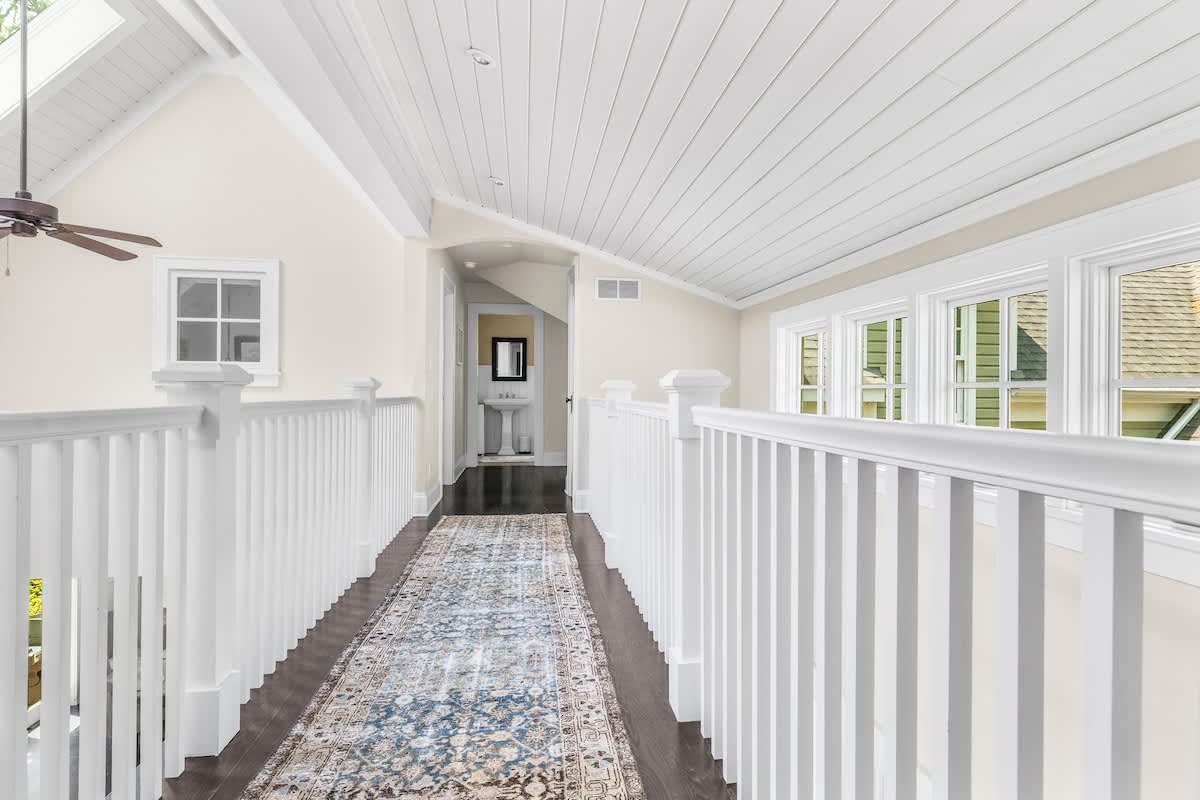 Long, bright hallway with dark wood floors, a patterned runner rug, white railings, and multiple windows, leading to a small bathroom with a pedestal sink at the far end.
