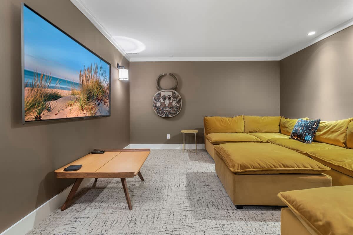Cozy media room with dark brown walls and patterned carpet, featuring a large mustard yellow sectional sofa, a wooden coffee table, and a wall-mounted TV for entertainment.