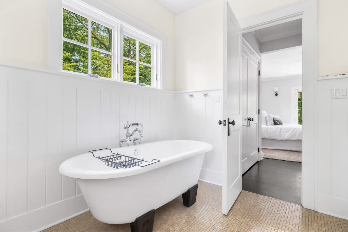A spacious bathroom featuring a white freestanding clawfoot tub with a bath caddy, set against white wainscoting and light walls, with a window overlooking greenery.