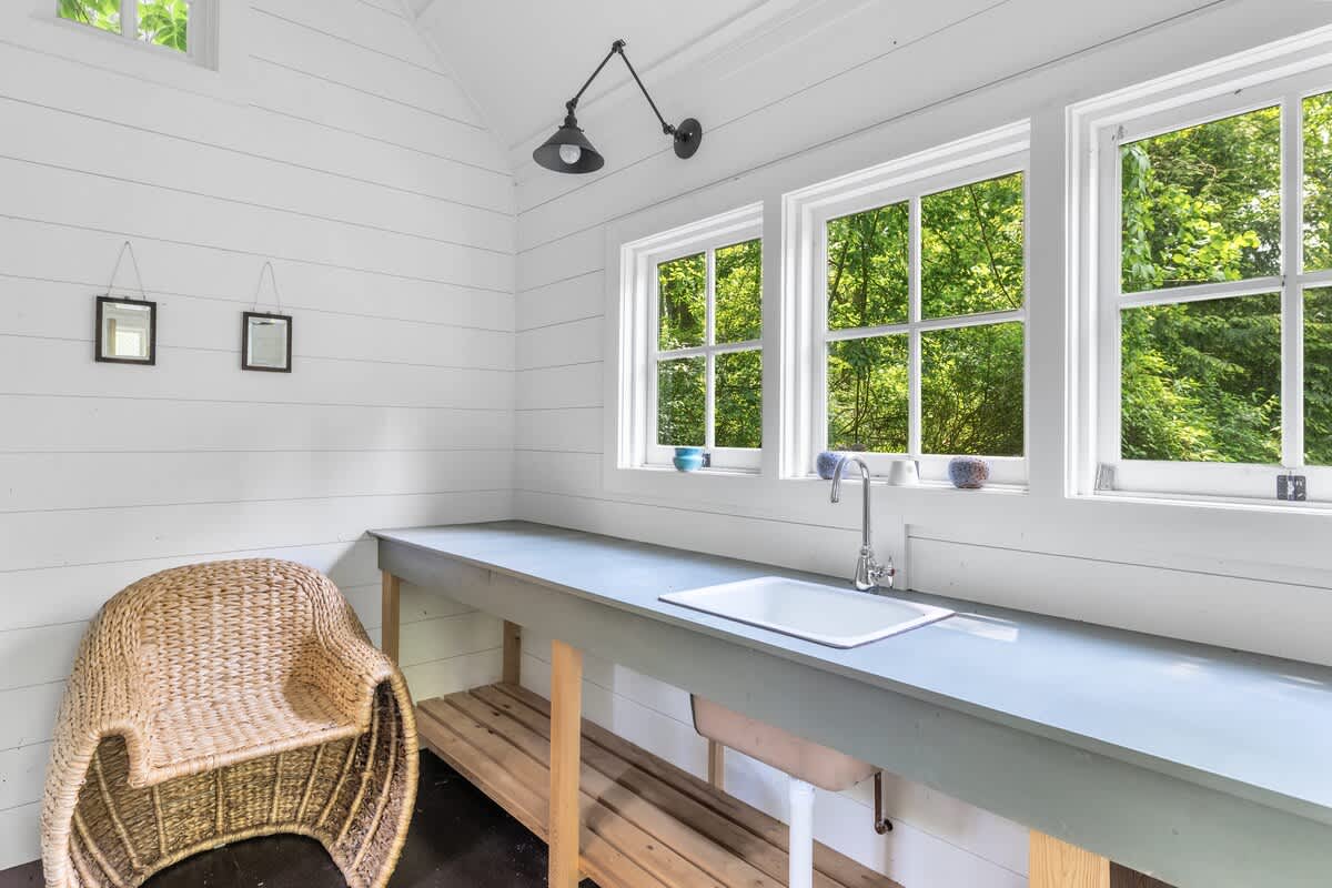 The interior of a bright shed, featuring white shiplap walls, a long workbench with an integrated sink, multiple windows offering views of greenery, and a wicker chair in the corner.
