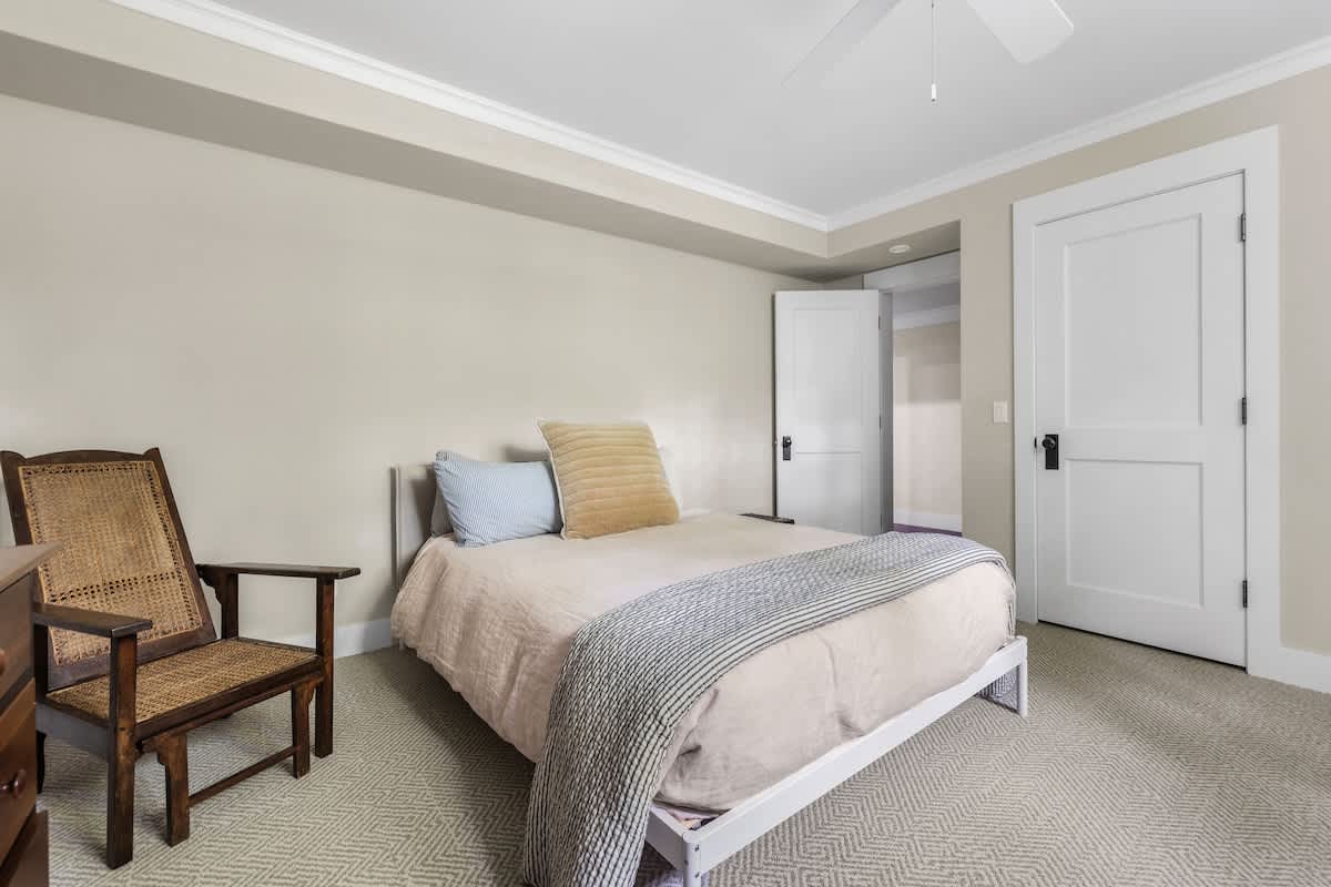 Neutral-toned bedroom with light beige walls and patterned carpet, featuring a bed with light bedding, a wooden chair, and white double doors leading to another room.