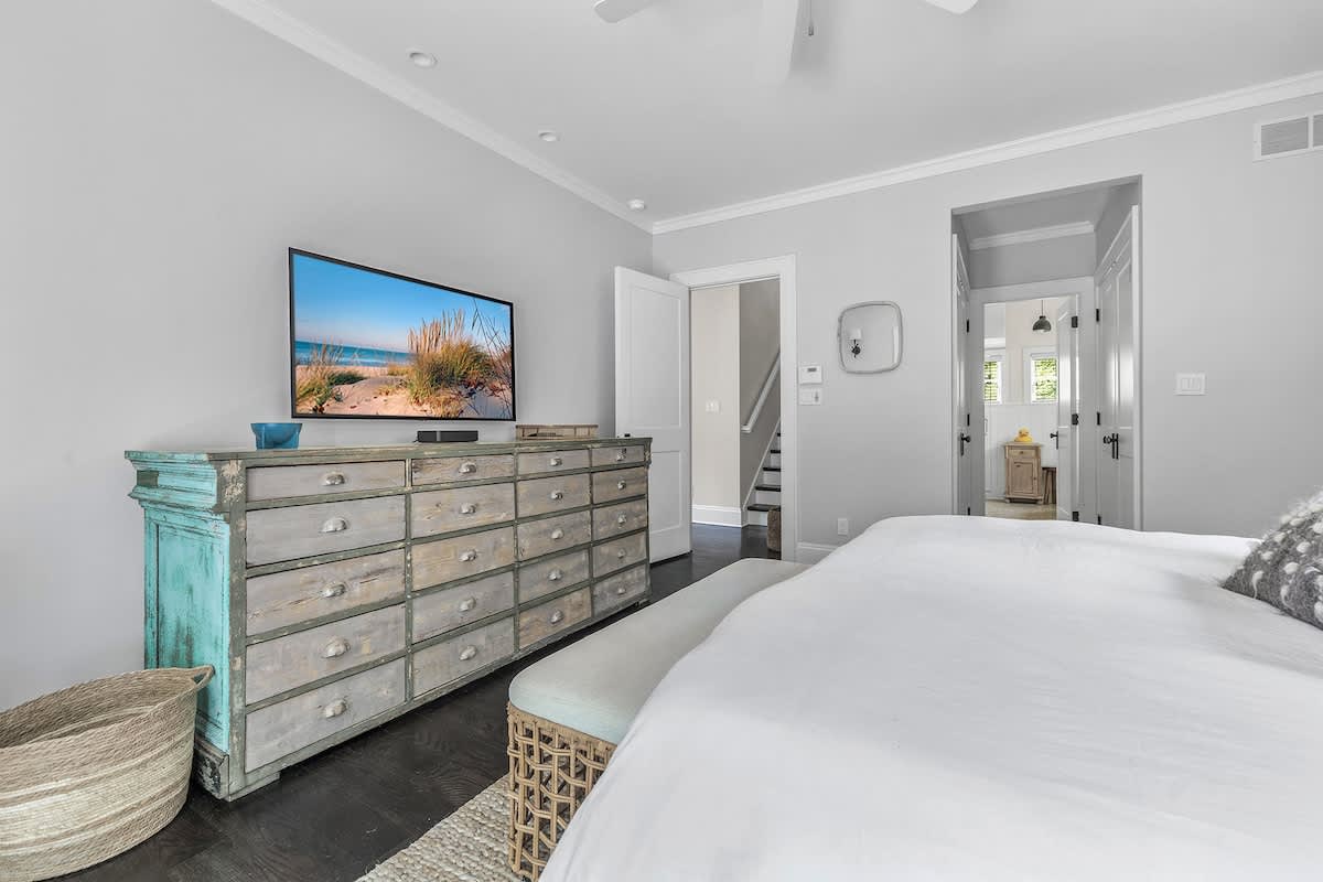A bright bedroom featuring a large distressed wooden dresser with a TV, a white bed with a light blue bench at its foot, and dark hardwood floors, leading to a hallway with stairs.
