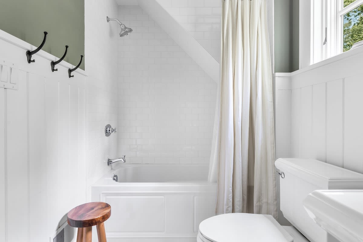 Compact bathroom with a white subway tile shower/tub combo, white wainscoting, and light green walls, featuring a shower curtain, toilet, and a small wooden stool.