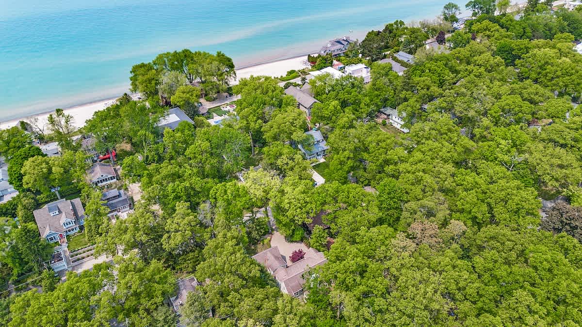 Wide aerial view showcasing the house nestled among dense green trees, with a sandy beach and a large turquoise body of water in the distance. Other houses are scattered throughout the wooded landscape.